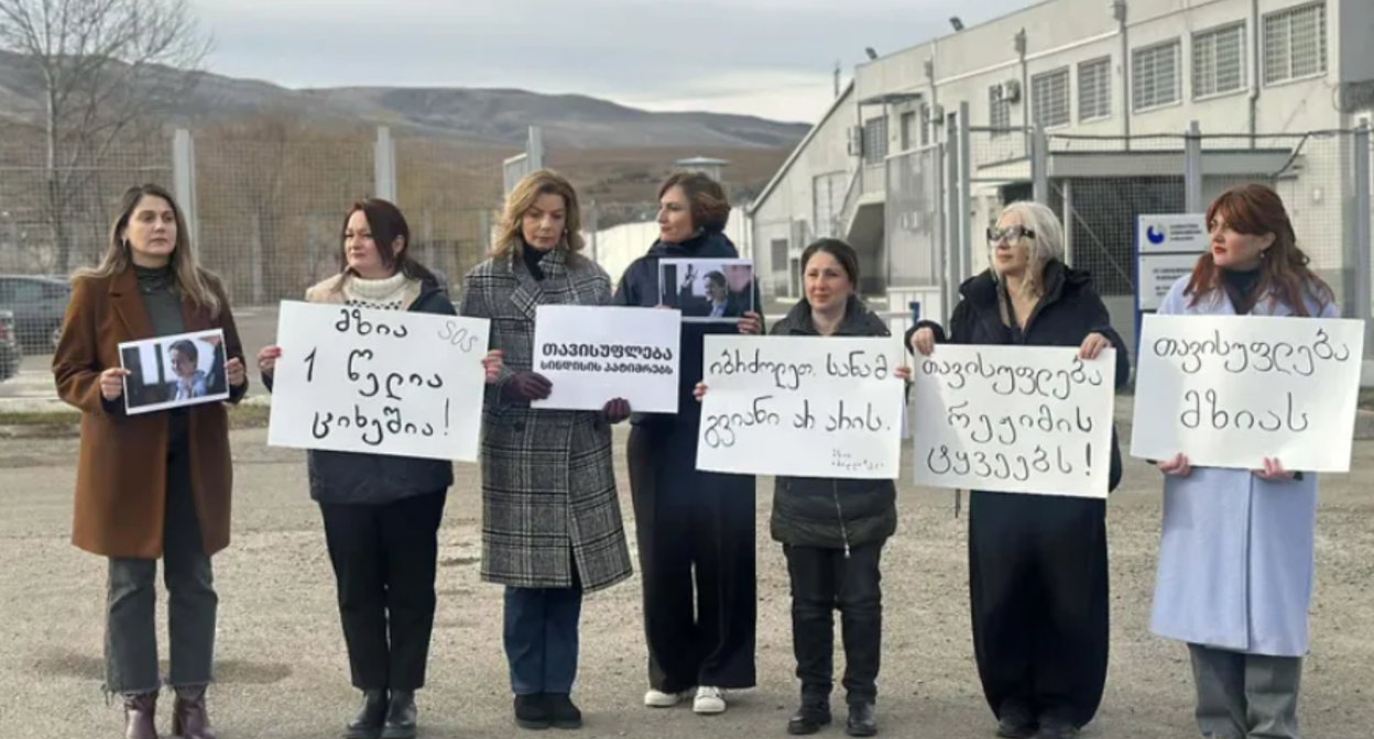 Participants of the action with posters in support of Mzia Amaglobeli. Screenshot of the photo "Interpressnews" from 11.01.26, https://www.interpressnews.ge/ru/article/172626-v-znak-solidarnosti-s-mziei-amaglobeli-zhenshchiny-politiki-sobralis-u-zdaniia-rustavskoi-zhenskoi-kolonii.