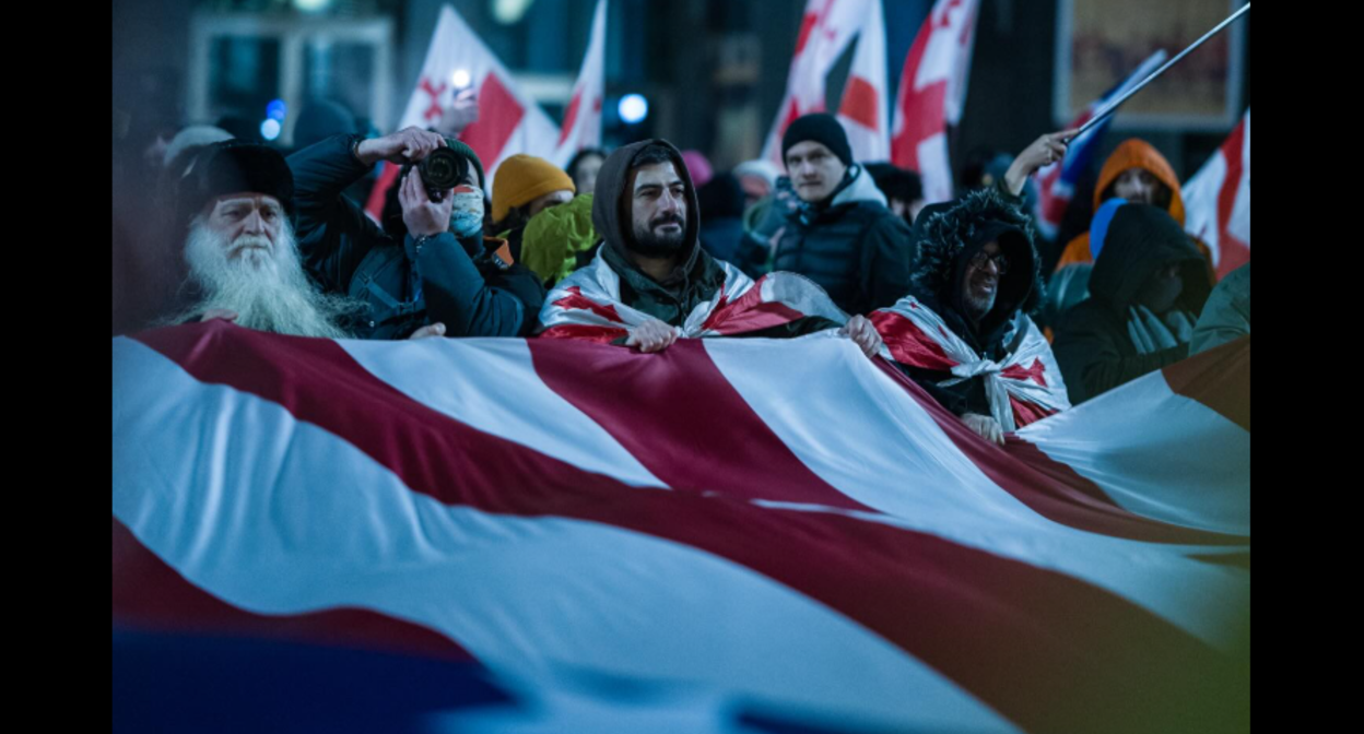 Protesters with a US flag. Screenshot of a photo by Mariam Qashbaia/Publika from February 28, 2026, https://www.facebook.com/photo/?fbid=1694177608648449&set=pcb.1694177725315104